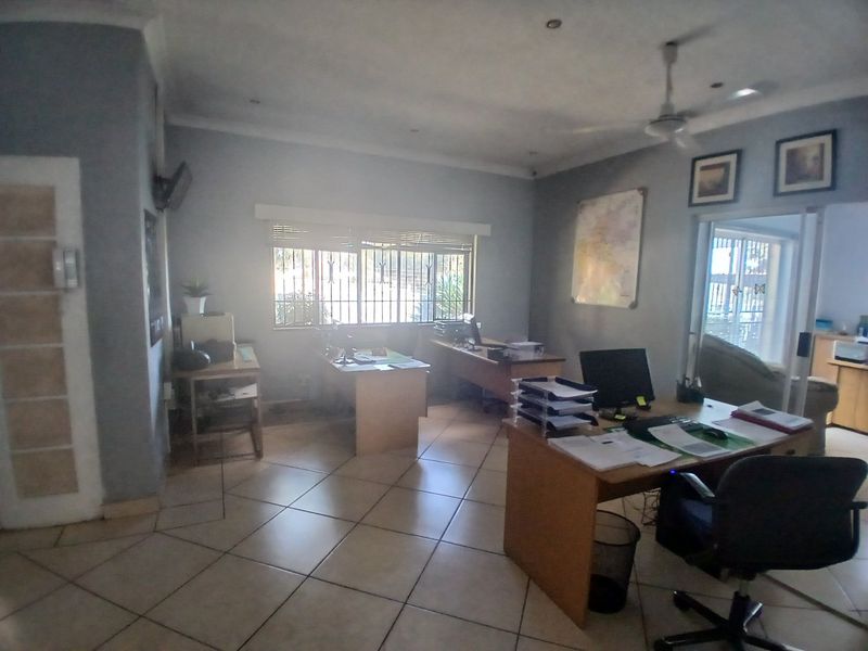 A functional commercial office space featuring tiled flooring, ceiling fan, and security bars on windows, providing a practical workspace environment.