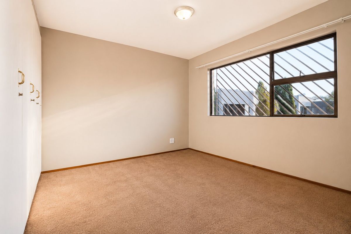 A bright, empty bedroom featuring neutral wall tones, fitted floor-to-ceiling wardrobes, and a window equipped with security bars.