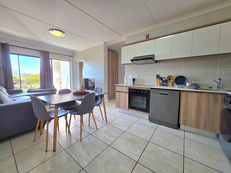 Modern open-plan living area featuring a contemporary kitchen with built-in cabinetry, integrated appliances, and tiled flooring, leading to a balcony with natural light.