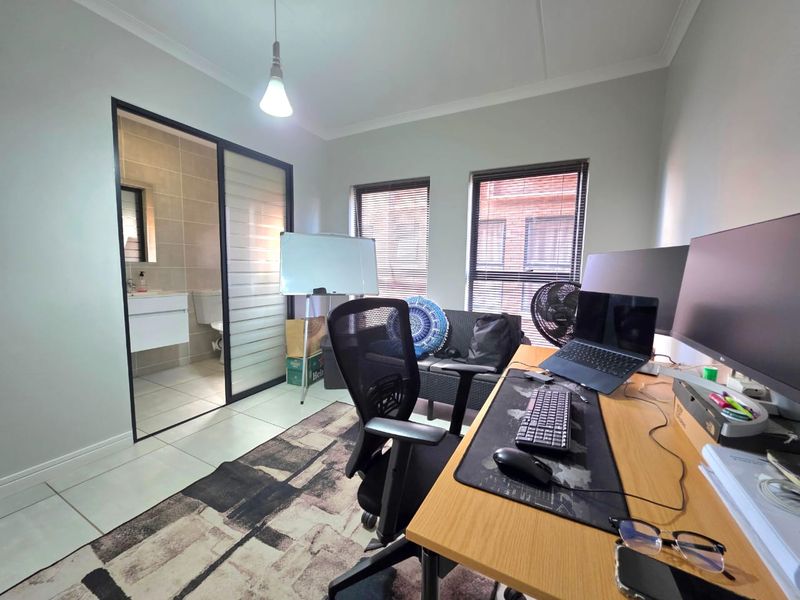 A functional home office space featuring tiled flooring, modern sliding glass door access to an en-suite bathroom, and large windows with venetian blinds.