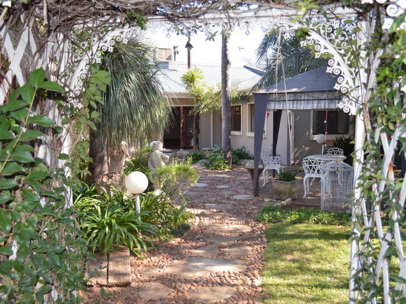 A charming rear garden featuring a stone pathway, mature landscaping, and a decorative metal archway leading to a covered patio area.