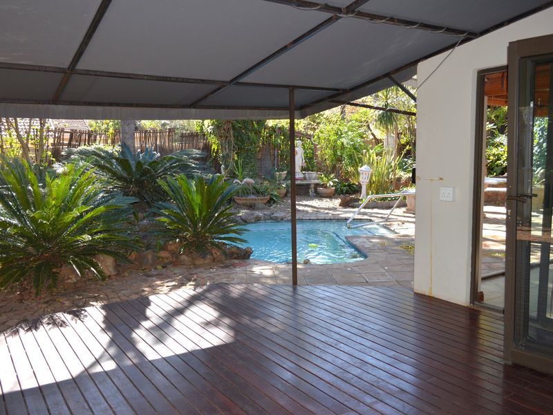 Outdoor entertainment area featuring a wooden deck, covered patio structure, and a swimming pool set within a landscaped garden.