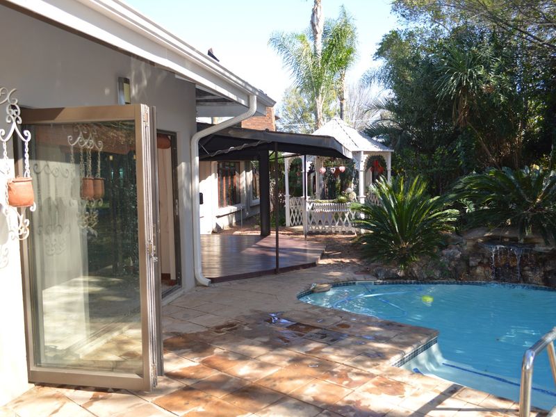 Rear exterior view featuring a swimming pool, stone patio, wooden deck, and covered outdoor entertainment area with bi-fold doors leading to the interior.