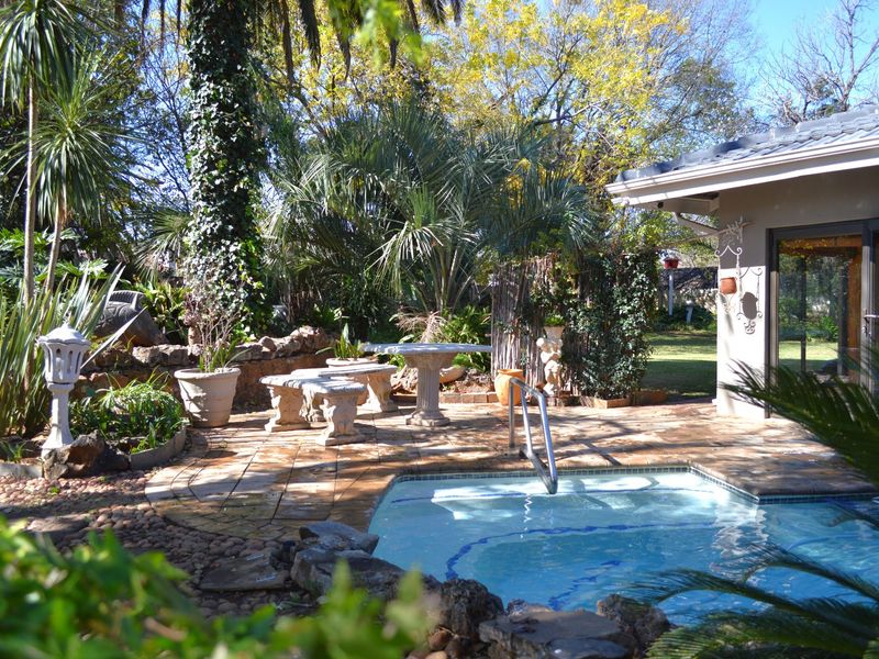 A well-maintained outdoor pool area featuring a tiled patio, stone landscaping, and a built-in pool with a handrail, set within a lush garden.