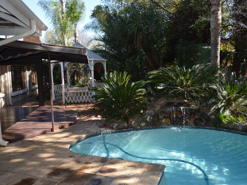 A private backyard pool area featuring a rock water feature, timber decking, and a charming gazebo set within a lush garden.