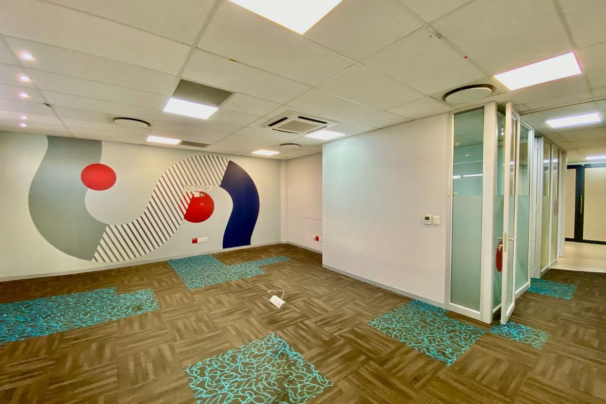 Empty commercial office space featuring a suspended ceiling, recessed lighting, glass partitioning, and patterned carpet tiles with some sections missing.