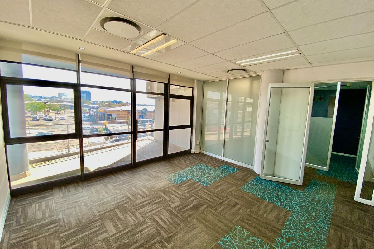 A bright, vacant commercial office space featuring floor-to-ceiling windows, glass partitions, suspended ceilings, and patterned carpet tiles.