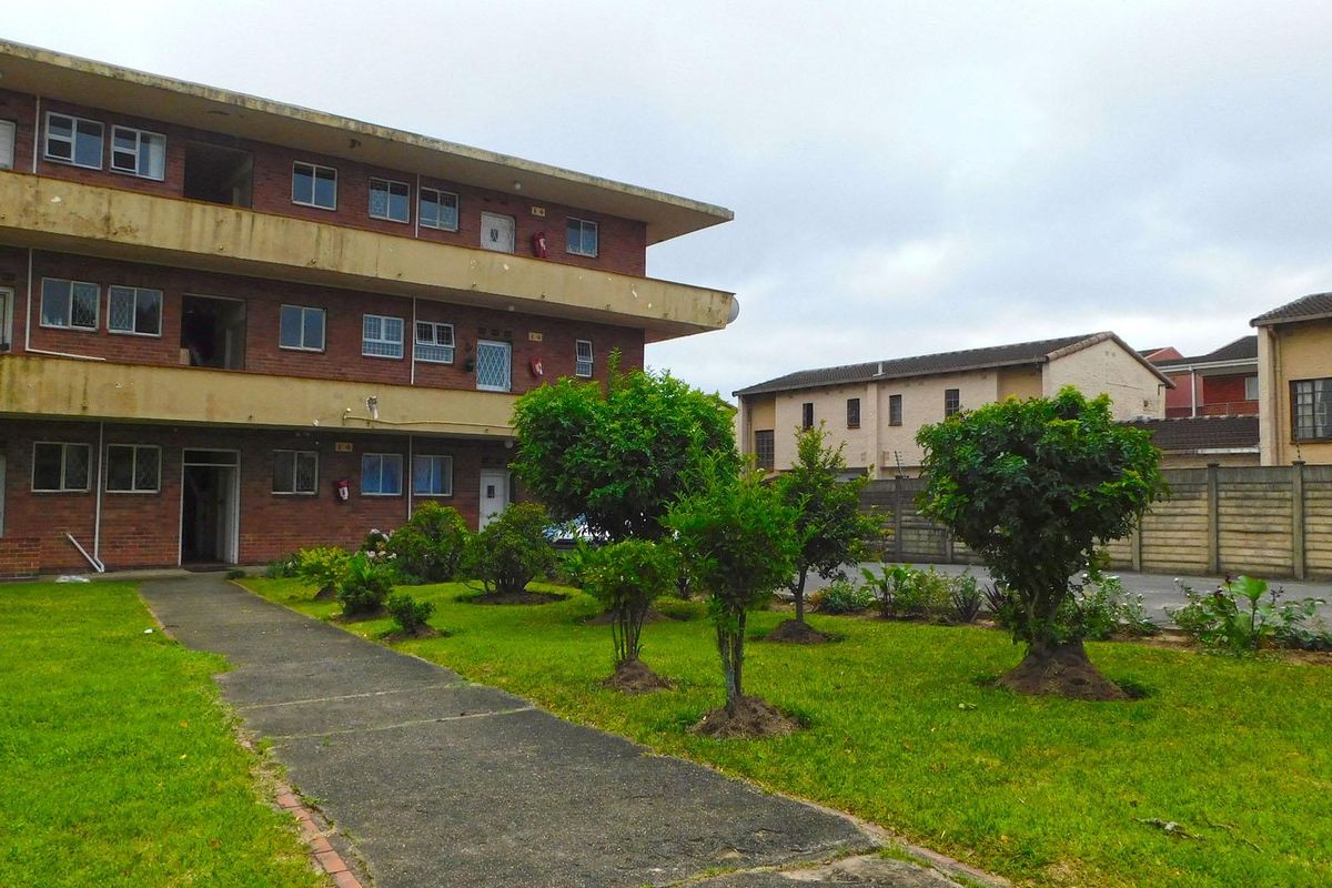 Exterior view of a multi-story brick apartment building featuring communal garden areas, concrete walkways, and a perimeter boundary wall.