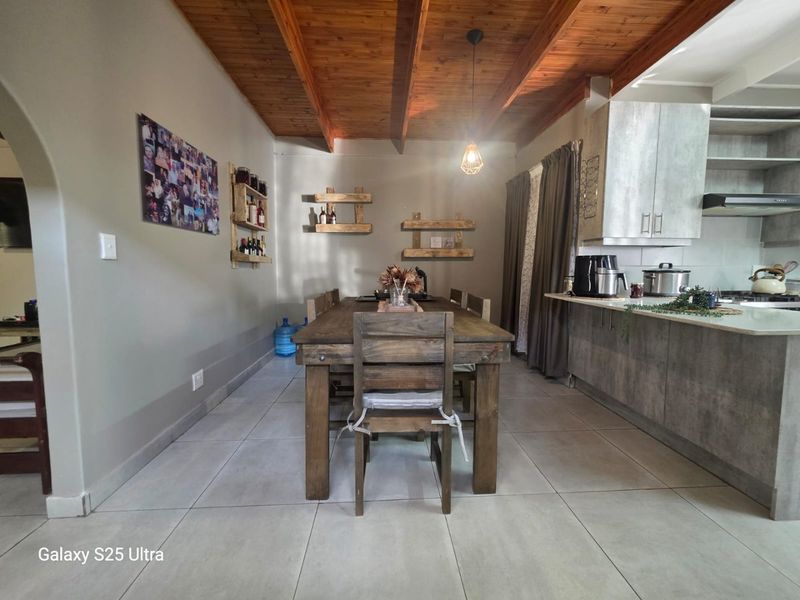 Open-plan kitchen and dining area featuring exposed timber beam ceilings, modern grey cabinetry, stone-look countertops, and neutral tiled flooring.