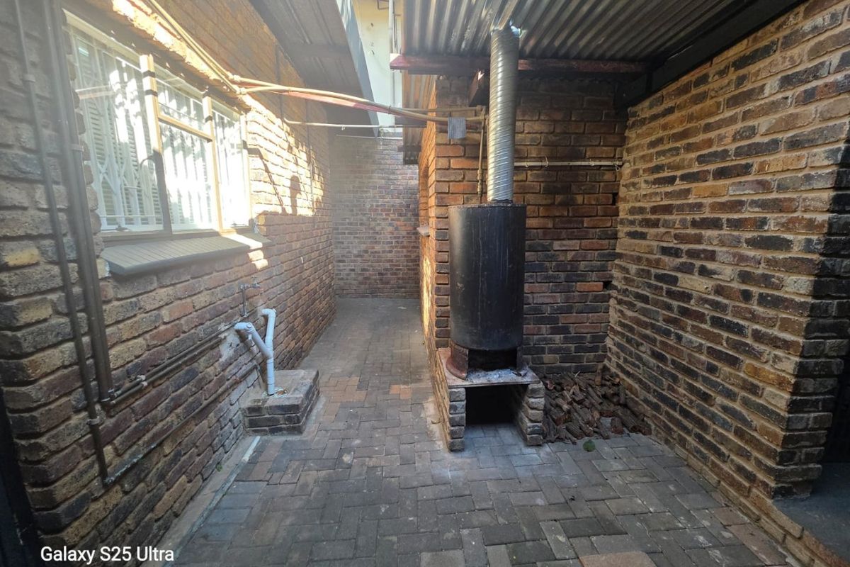 A narrow exterior rear service area featuring brick paving, exposed brick walls, and a built-in outdoor fireplace with a metal chimney flue.