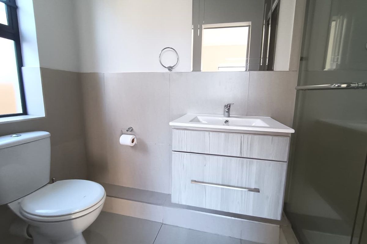 A modern, clean bathroom featuring a vanity cabinet with a basin, a toilet, and a glass-enclosed shower with neutral wall and floor tiling.