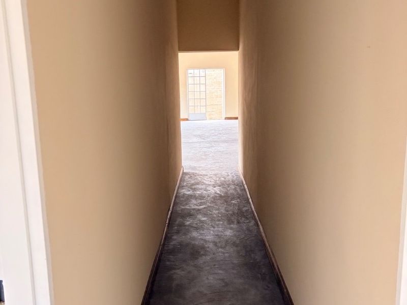 A narrow, empty hallway featuring concrete flooring and neutral wall finishes, leading towards an open living space with a glass door.