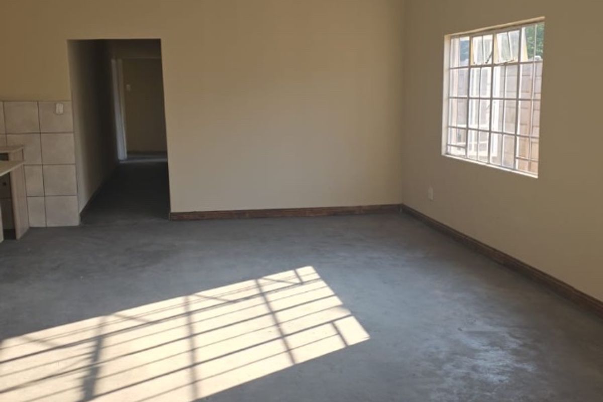 An empty, open-plan living area featuring neutral walls, security-barred windows, and unfinished concrete flooring, offering a blank canvas for renovation.