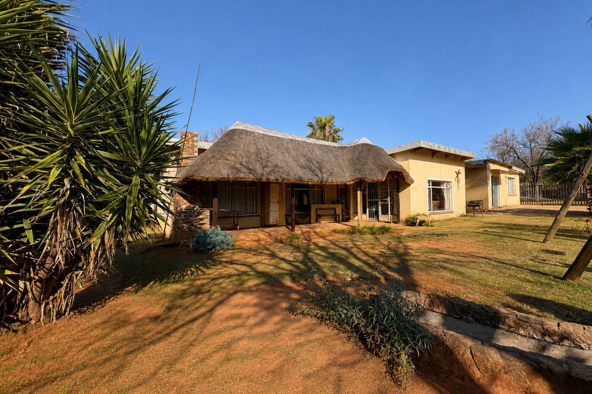 Rear view of a single-story house featuring a traditional thatch roof, covered patio area, and surrounding garden space with natural terrain.