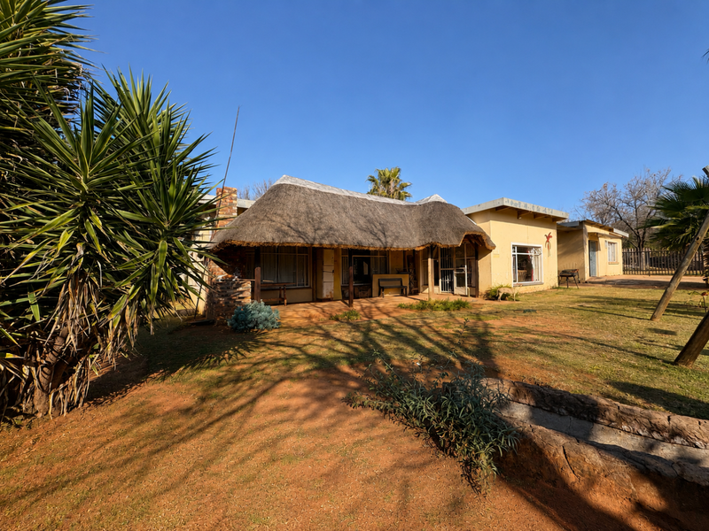Rear view of a single-story house featuring a traditional thatch roof, covered patio area, and surrounding garden space with natural terrain.