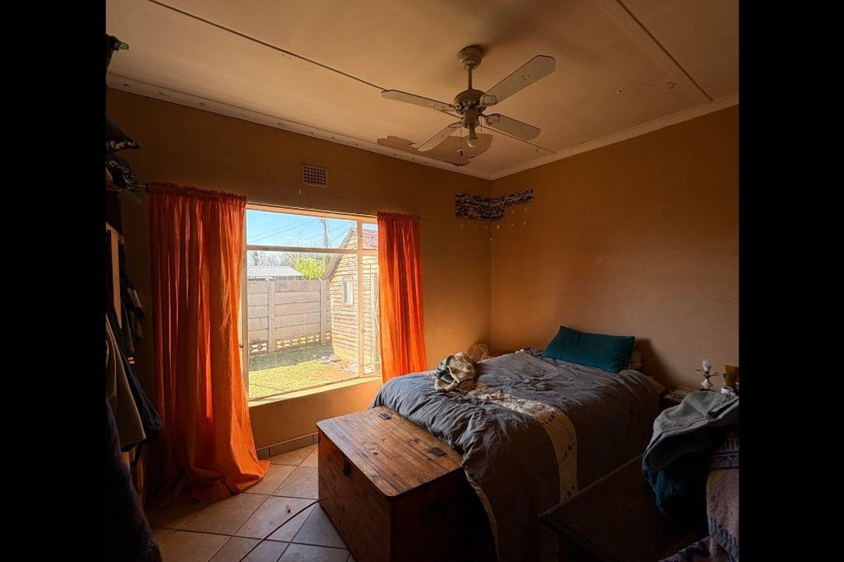 A bedroom featuring tiled flooring, a ceiling fan, and a large window with natural light. The ceiling requires repair due to visible surface damage.