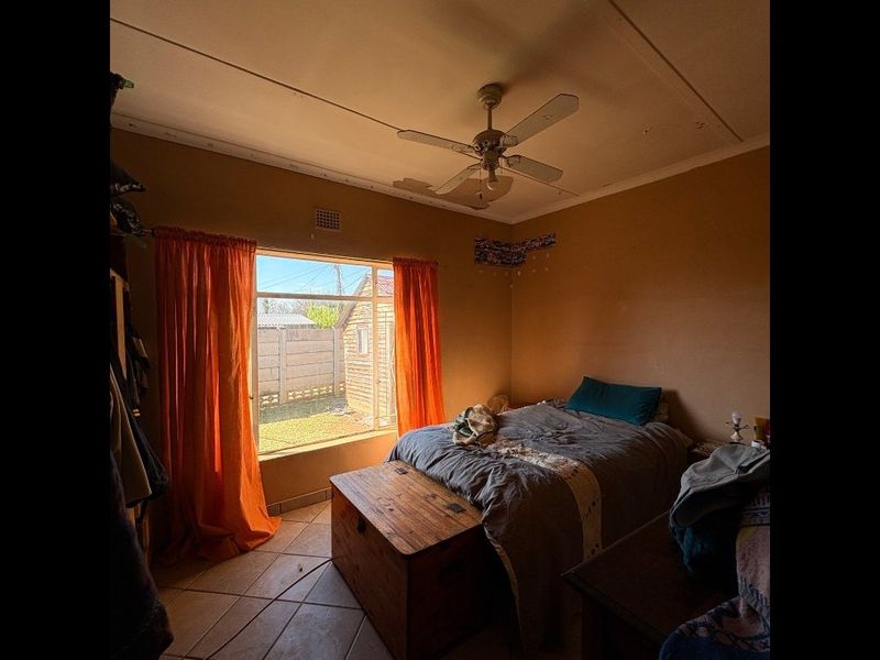 A bedroom featuring tiled flooring, a ceiling fan, and a large window with natural light. The ceiling requires repair due to visible surface damage.