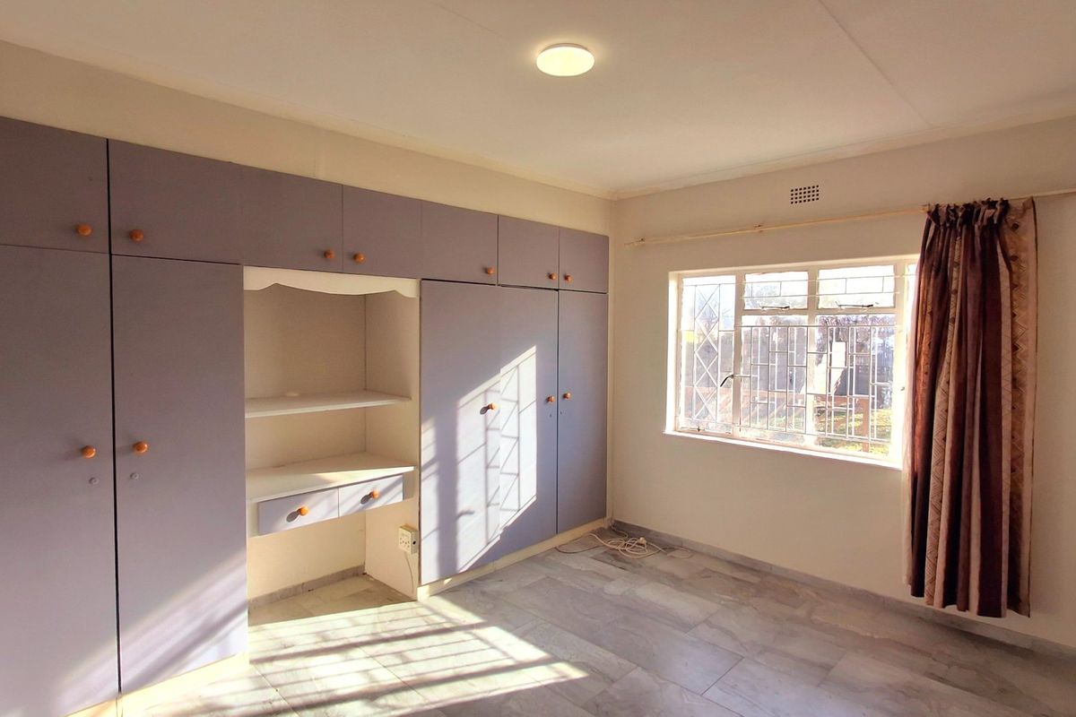 A bright, empty bedroom featuring extensive built-in cupboards, tiled flooring, and security bars on the window.