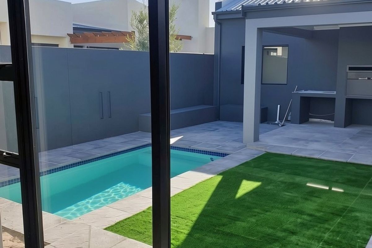 Modern rear courtyard featuring a swimming pool, paved patio area, and a built-in braai, viewed through large sliding glass doors.