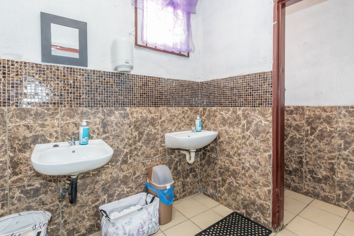 A functional commercial bathroom featuring dual wall-mounted basins, marble-effect wall tiling, and mosaic accents, requiring some maintenance and cosmetic updates.