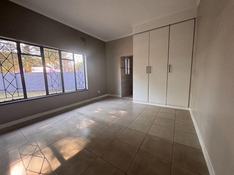 A bright, empty bedroom featuring neutral tiled flooring, built-in cupboards, and large windows with security bars, offering a clean and versatile living space.