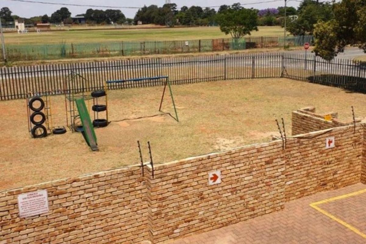 An outdoor area featuring a brick boundary wall with electric fencing, paved driveway space, and a grassed yard with playground equipment.
