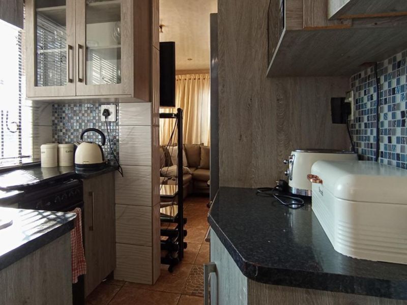 Compact kitchen featuring wood-look laminate cabinetry, mosaic tile backsplash, and dark countertops, with a direct view into the adjacent living area.