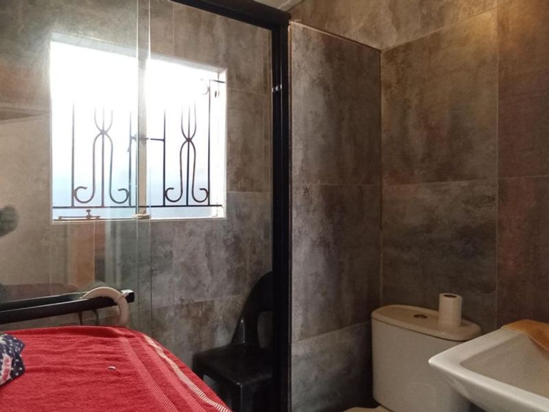 A functional bathroom featuring a shower enclosure, toilet, and washbasin with modern grey wall tiling and security bars on the window.