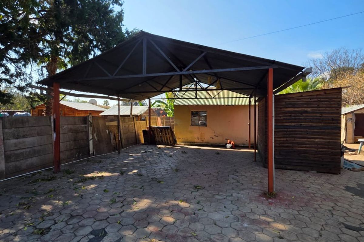 An outdoor rear area featuring a metal carport structure over a paved surface, enclosed by precast concrete walls and timber screening.