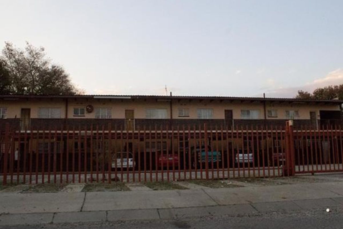 Front elevation of a multi-story apartment complex featuring a secure perimeter fence and covered ground-level parking bays.