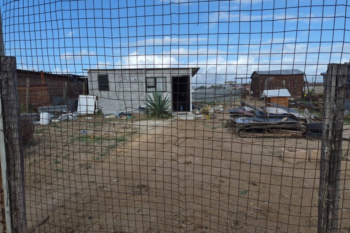 View of a residential plot featuring a basic concrete structure, wire fencing, and scattered debris on the grounds.