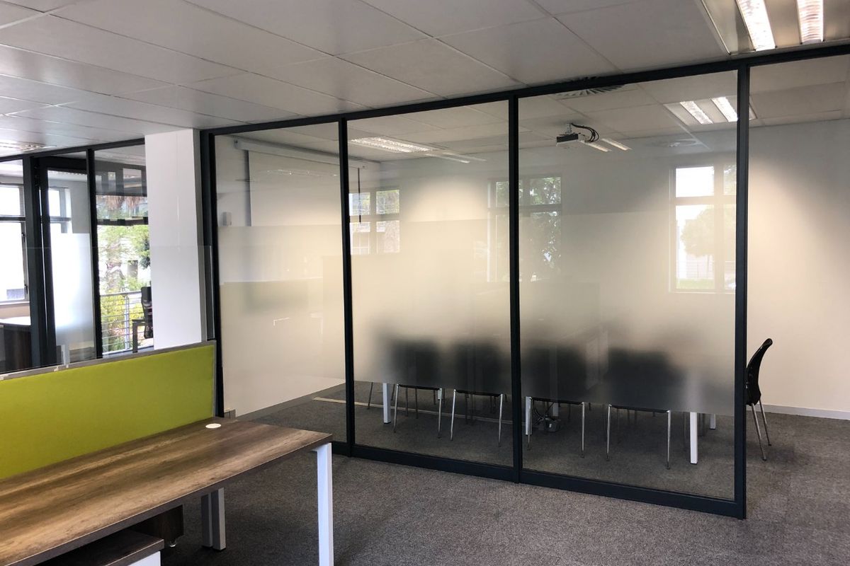 Modern commercial office space featuring a glass-partitioned meeting room with frosted privacy film, suspended ceiling, and carpeted flooring.