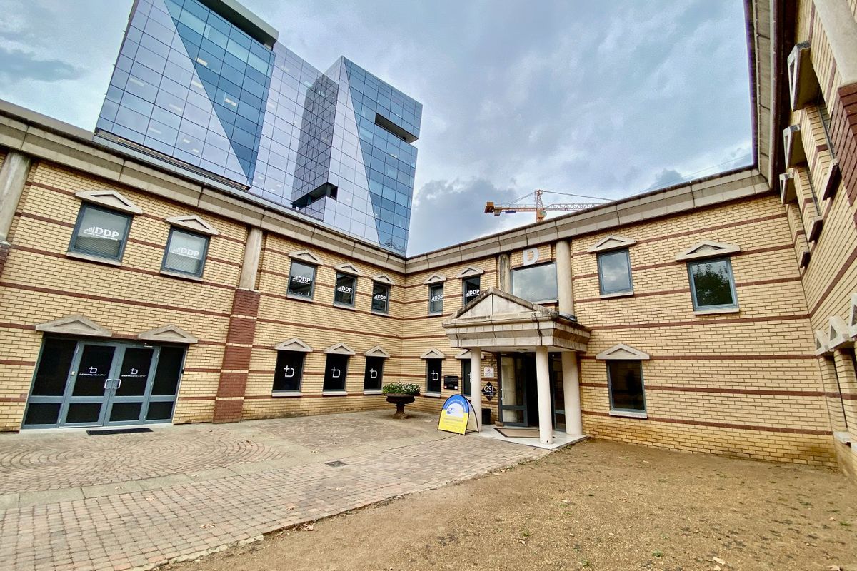 A commercial courtyard area featuring brick facade architecture, a paved entrance approach, and a modern glass office building in the background.