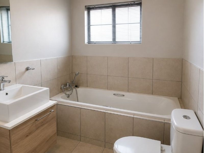 A clean, modern bathroom featuring a built-in vanity with basin, a tiled bathtub with handheld shower, and a standard toilet.