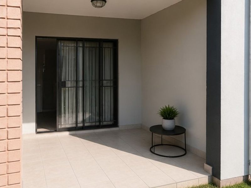 A clean, covered patio area featuring tiled flooring, a sliding security gate, and a brick pillar, offering a functional outdoor space.