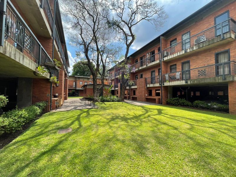 A well-maintained communal garden area between apartment blocks, featuring brick facades, metal balcony railings, and paved walkways with mature trees.