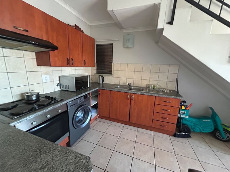 Functional kitchen featuring wood-finish cabinetry, tiled splashbacks, integrated appliances, and a convenient layout situated beneath an internal staircase.