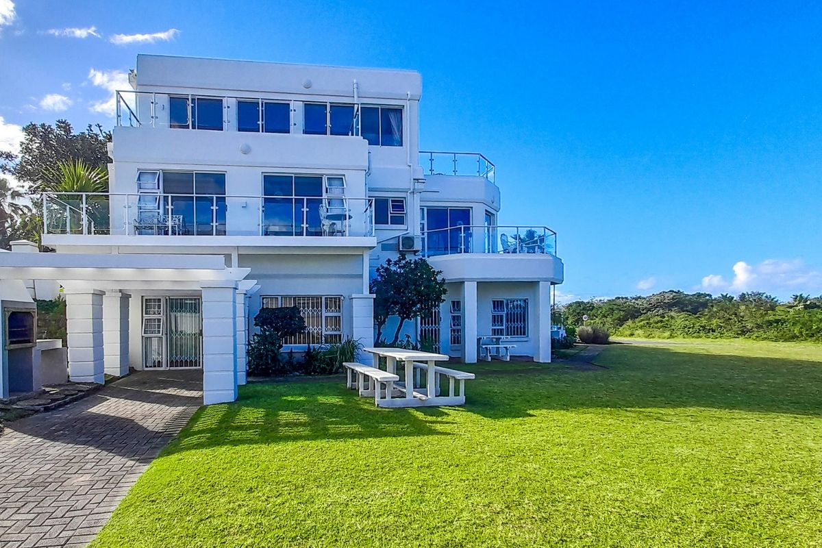 A multi-level residential exterior featuring a paved patio, built-in braai, and upper-level balconies with glass balustrades overlooking a spacious lawn.