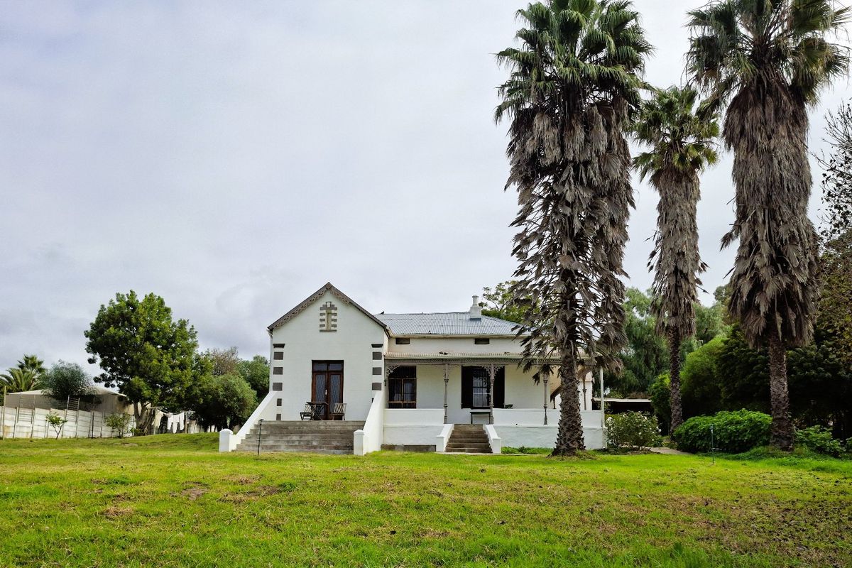 A charming historic-style house featuring a covered veranda, concrete stairs, and a corrugated metal roof, set within a spacious garden with mature palm trees.
