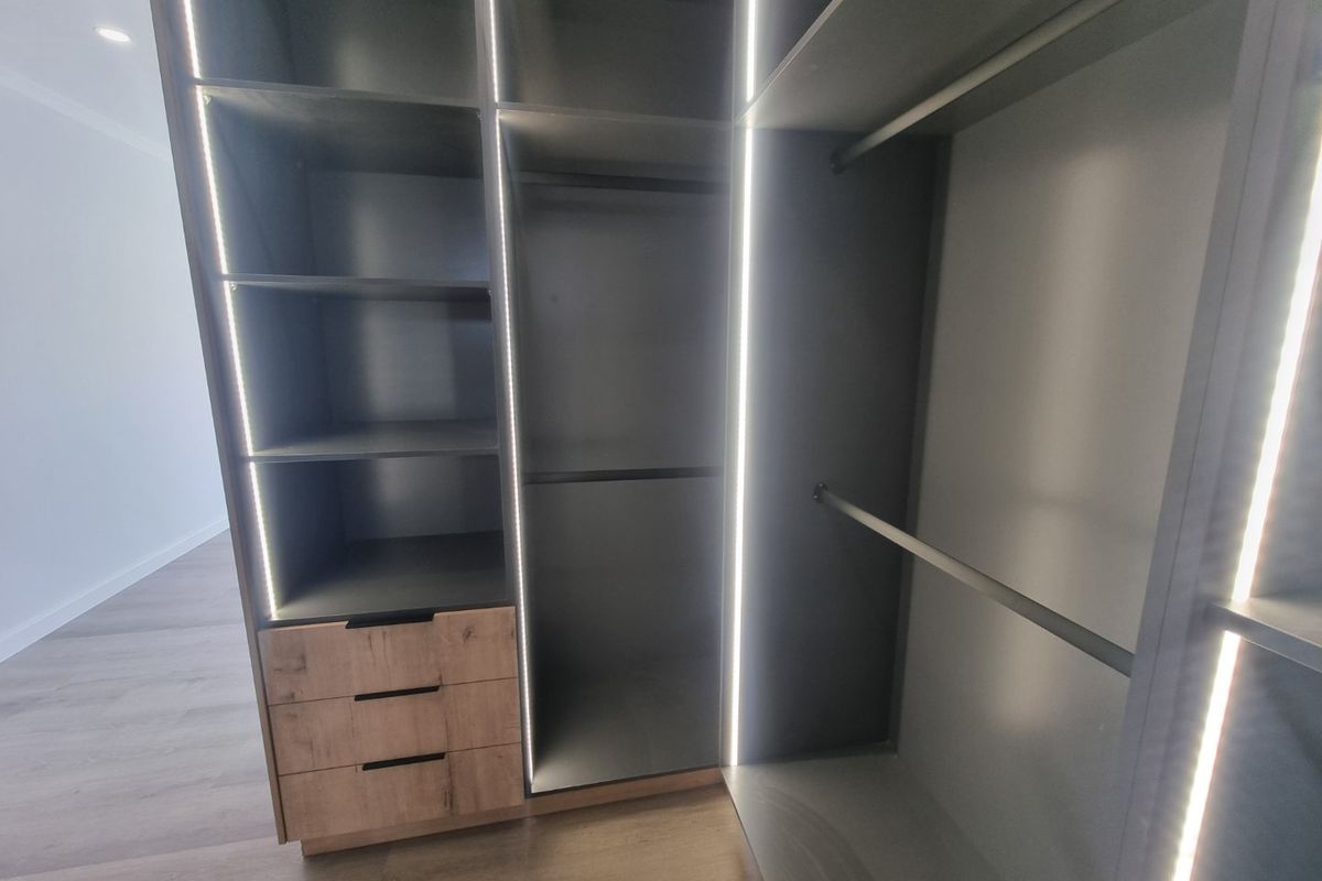 Modern built-in wardrobe featuring integrated LED lighting, open shelving, and wooden drawer units, set within a bedroom with laminate flooring.