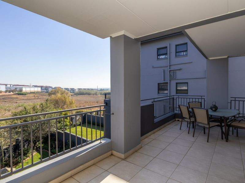 A spacious covered balcony featuring tiled flooring and a metal balustrade, offering an open view of the surrounding landscape and neighboring complex.
