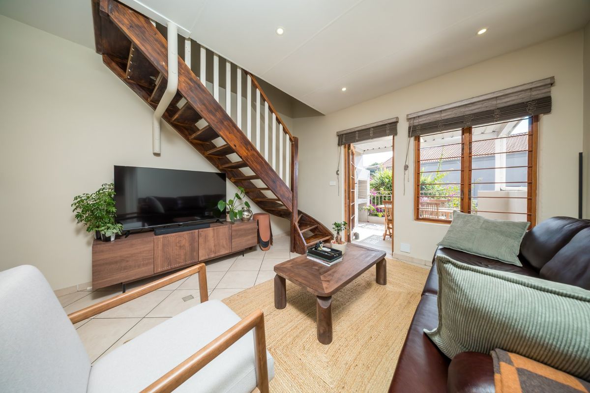 A bright living room featuring a prominent wooden staircase, tiled flooring, and direct access to an outdoor patio area through a timber door.