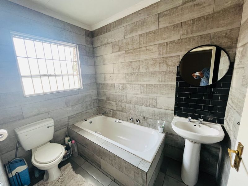 A functional bathroom featuring a built-in bathtub, pedestal basin, toilet, and modern wood-look wall tiling with a security-barred window.