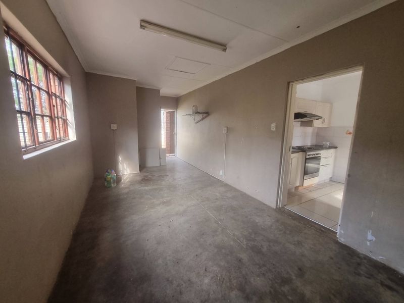 An empty living area with concrete flooring and steel-framed windows, leading into a separate kitchen space.