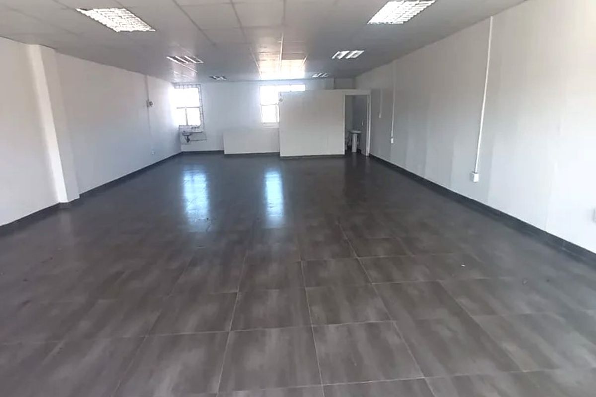 A spacious, empty commercial office area featuring tiled flooring, a suspended ceiling with recessed lighting, and a small partitioned section.