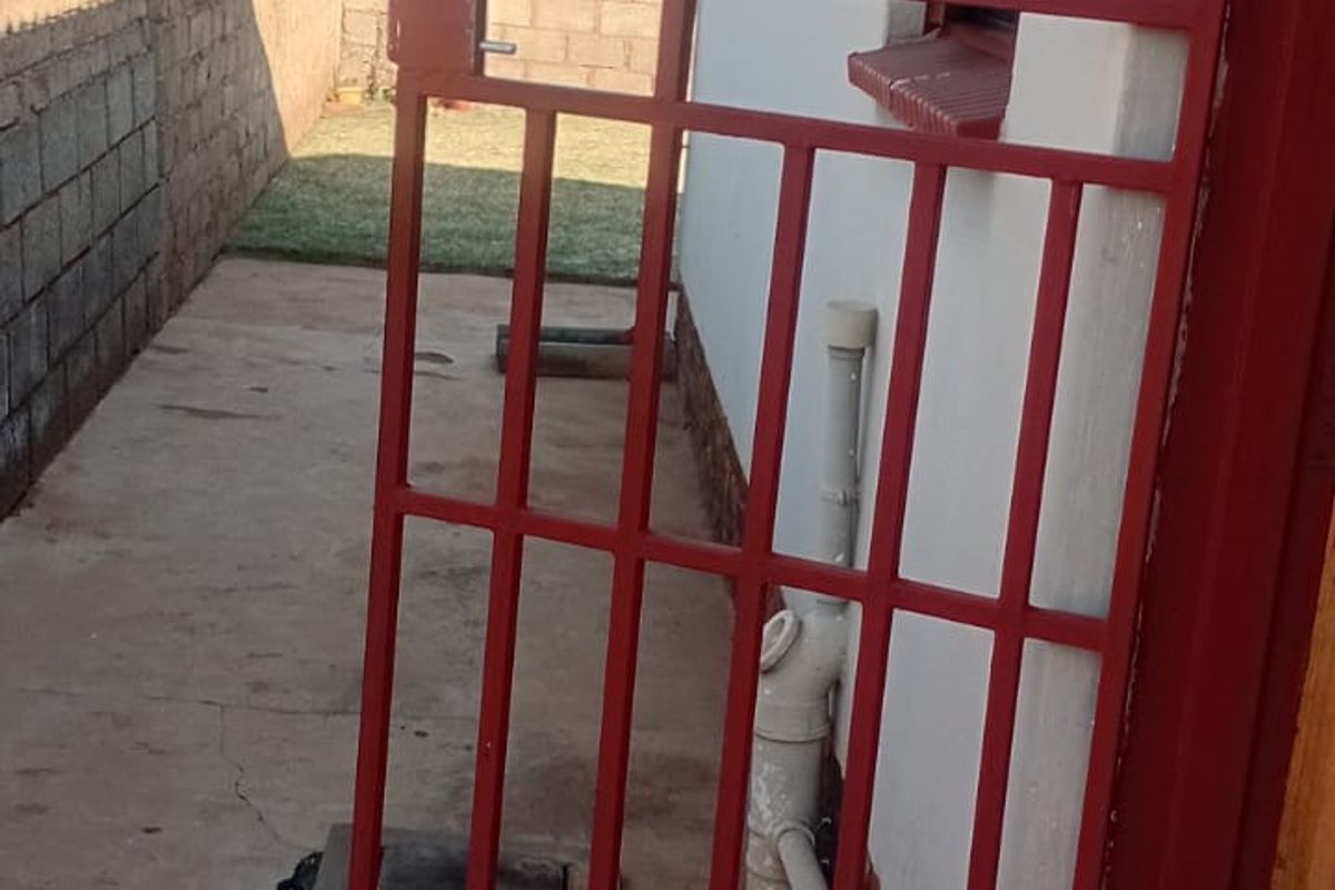 A side exterior view showing a red metal security gate, concrete paving, and a boundary wall along the side of the house.