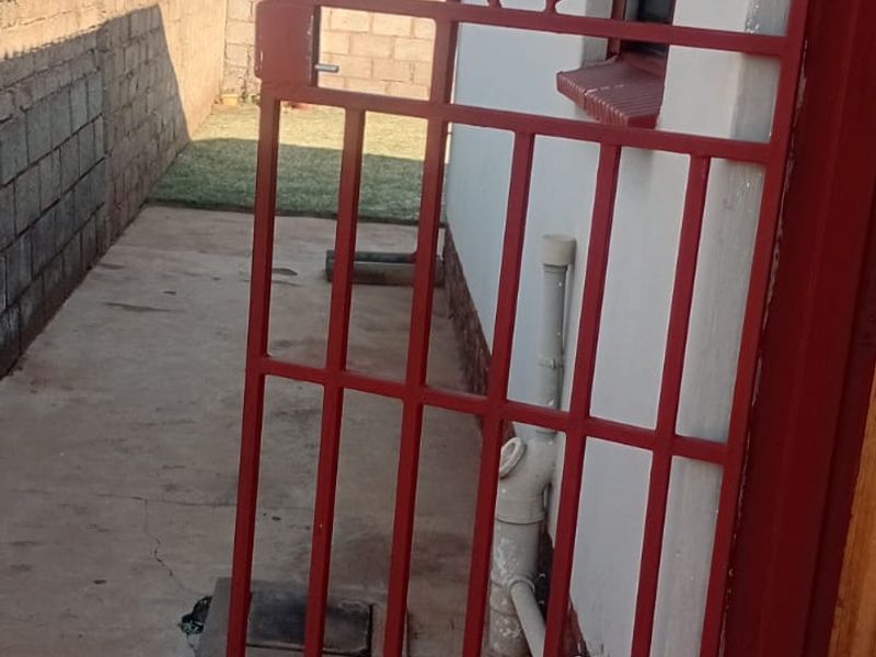 A side exterior view showing a red metal security gate, concrete paving, and a boundary wall along the side of the house.