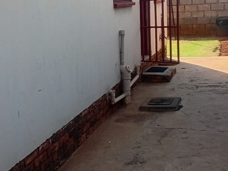 Rear exterior view of the property showing a concrete pathway, security gate, and exposed brick foundation with visible wear.
