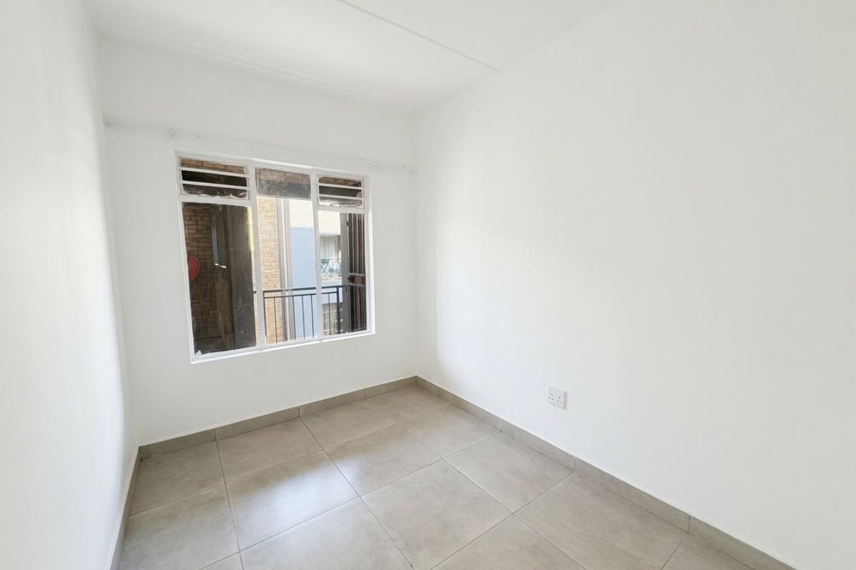 A bright, empty bedroom featuring neutral tiled flooring, white walls, and an aluminium-framed window providing natural light.