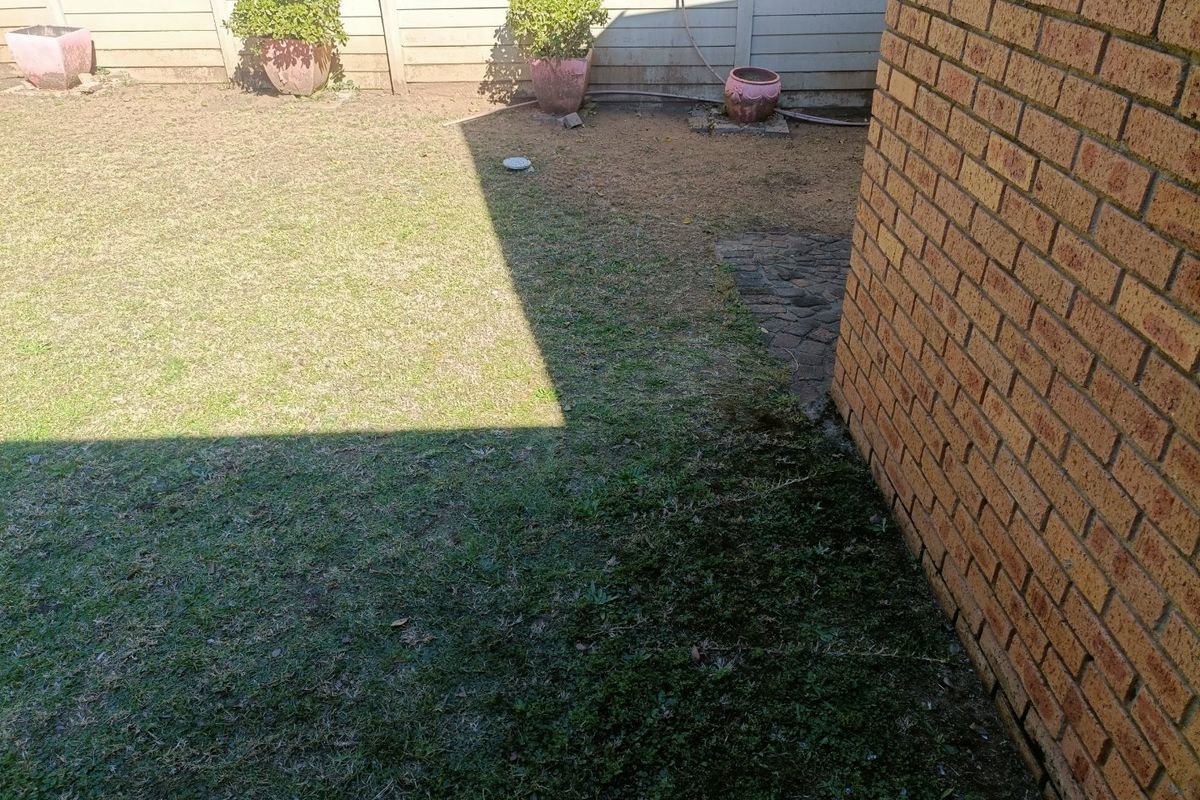 A rear garden area featuring a brick exterior wall, precast concrete boundary fencing, and a lawn requiring maintenance.
